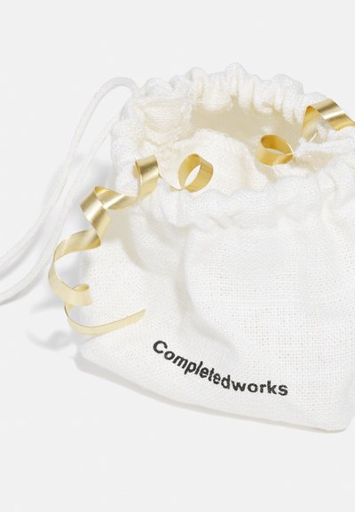 Sac en coton à cordon de serrage blanc avec une finition texturée, présentant des accents en ruban doré et "Completedworks" imprimé en noir.