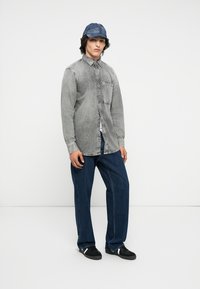 Grijze denimoverhemd met een voorkoker, gecombineerd met donkerblauwe wijdbeinige jeans. Het model draagt een blauwe pet met patroon en zwarte sneakers.