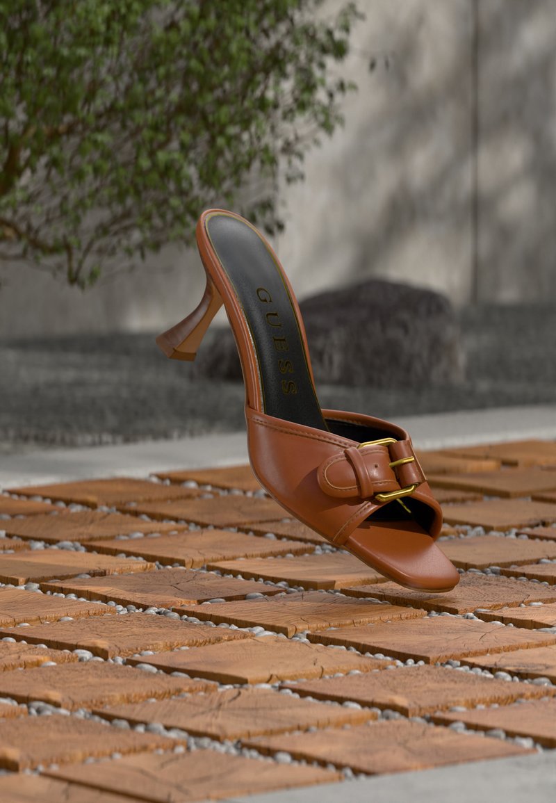 Mule à talon haut en cuir marron avec détail boucle, marquée « Guess » sur la semelle intérieure, posée sur des carreaux en bois à motifs avec de la verdure et un mur en béton en arrière-plan.