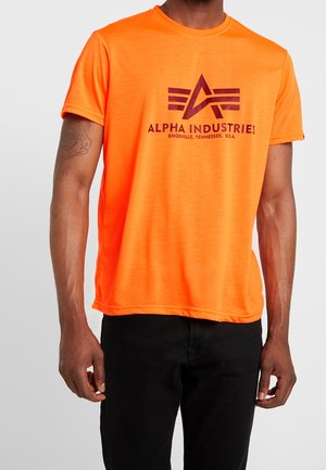 Homme portant un t-shirt manche courte orange vif avec le logo et le texte Alpha Industries en rouge foncé, associé à un pantalon noir.