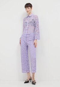 Chemisier en dentelle lilas avec col, doté de longues manches transparentes, associé à un pantalon en dentelle assorti. Les deux présentent un motif floral et une coupe courte.