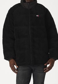 Czarna polarowa kurtka z wysokim kołnierzem, zamkiem błyskawicznym i elastycznymi mankietami. Posiada małą naszywkę z logo na klatce piersiowej oraz pikowaną fakturę.