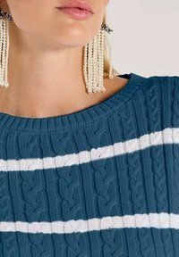 Nahaufnahme einer Person, die Perlenfransenohrringe und einen blauen Strickpullover mit weißen horizontalen Streifen trägt.