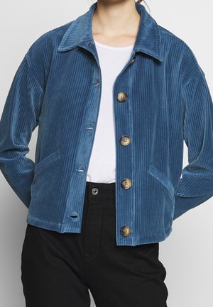 Vrouw draagt een blauw corduroy jasje met knopen over een wit shirt en zwarte broek, handen in de zakken van het jasje gestopt.