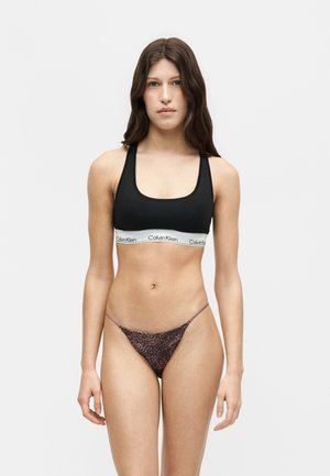 Vrouw met lang bruin haar, gekleed in een zwarte Calvin Klein sportbeha en bikinibroekje met luipaardprint, staand tegen een effen lichte achtergrond.