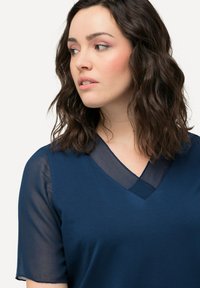 Námořnický top s výstřihem do V, krátkými průhlednými rukávy a hladkou texturou. Tkanina působí lehce a poloprůhledně na ramenou.