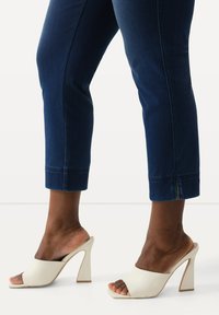 Mules de tacón abiertos en blanco con un acabado suave y un tacón en bloque esculpido, combinados con jeans oscuros de longitud hasta el tobillo con dobladillo. 