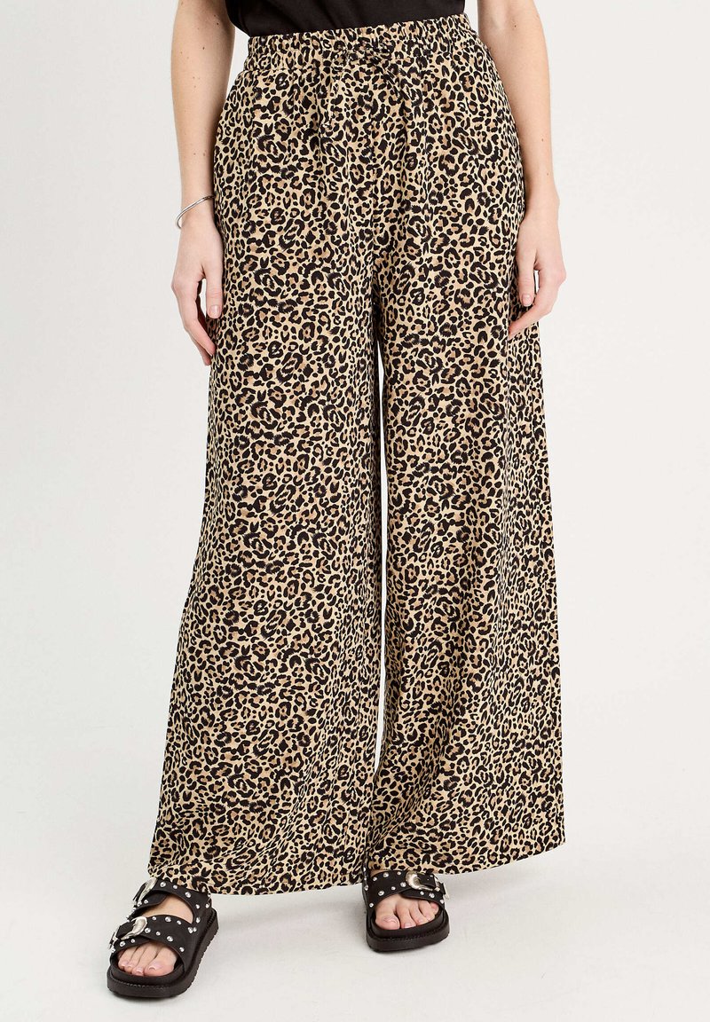 Leopardmønstret, vide bukser har en elastisk talje, en beige baggrund og markante sorte pletter. Stylet med sorte sandaler.
