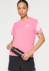 Ροζ αθλητική φανέλα με λευκό λογότυπο και τρεις ρίγες στα μανίκια, σε συνδυασμό με μαύρο σορτς και μαύρο παγούρι.