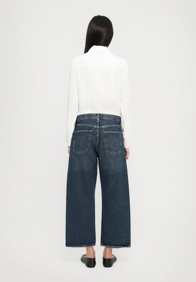 Wide-leg dark blue jeans with a high waist and faded texture, paired with a long-sleeve white blouse featuring a collar and back panel.