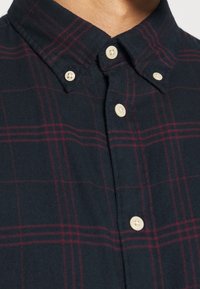 Mörkblå skjorta med knappar och röd rutmönstring, med en knapp-ner krage och krämfärgade knappar, som bärs på en persons övre bröst.