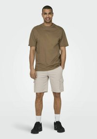 Brun kortærmet t-shirt, beige cargo-shorts, sorte sneakers og hvide sokker. Stående mandlig model i afslappet tøj, med hænderne i lommerne.