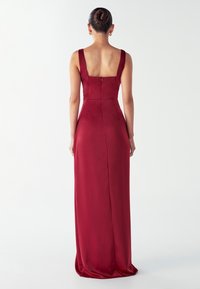 Ženska z lasmi v banana pričeski, oblečena v brezrokavno temno rdečo obleko do tal z kvadratnim izrezom, stoji pred enobarvno belo ozadjem.