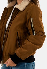 Giacca bomber marrone con design trapuntato, colletto in pelliccia sintetica, polsini a coste neri e tasche laterali. Presenta una tasca con zip sulla manica.