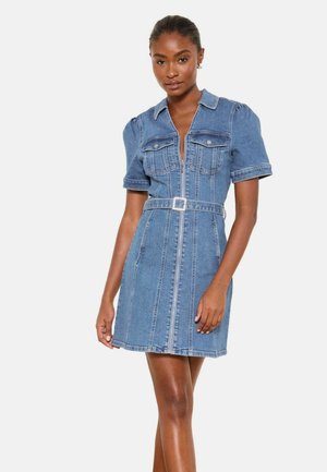Frau trägt ein blaues Denimkleid mit kurzen Puffärmeln, vorderem Reißverschluss, Brusttaschen und passendem Gürtel in der Taille, steht vor weißem Hintergrund.