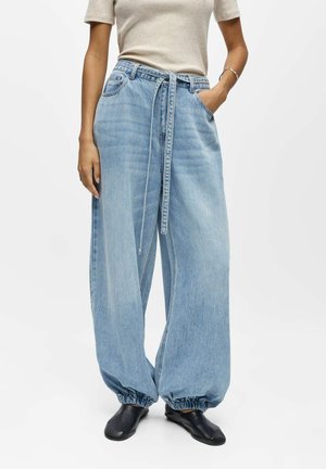 Personne portant un jean bleu clair à jambes larges avec une ceinture, des poignets élastiques, un haut beige à manches courtes et des chaussures noires à enfiler.