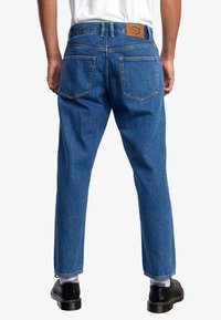 Pantalones vaqueros de mezclilla en azul oscuro, con corte recto, que cuentan con un parche trasero de cuero marrón y dos bolsillos traseros con costuras en contraste.