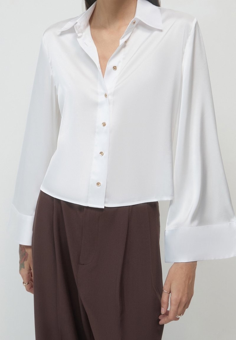 Personne portant un chemisier blanc en satin à manches longues avec des boutons dorés et un pantalon marron taille haute, montrée des épaules jusqu’au milieu des cuisses.