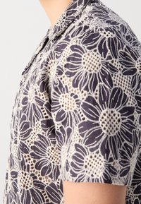 Chemise à manches courtes avec fond bleu marine et grands motifs en dentelle florale beige, avec un col pointu et des détails découpés complexes.