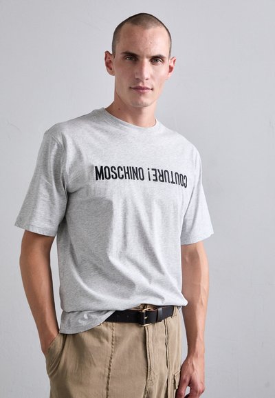 Hallid lühikeste varrukatega hall T-särk, mille ees on must "MOSCHINO" ja "COUTURE" trükk. Pehme, puuvillane materjal klassikalise lõikega.