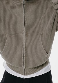 Sweat à capuche zippé en maille beige doux avec texture côtelée, comprenant deux poches avant et un chemise rayée blanche superposée dessous.