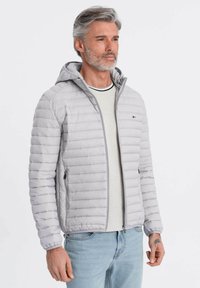 Lys grå pufferjakke med hætte, med en quiltet struktur og sidelommer, båret over en lys grå skjorte og blå jeans.