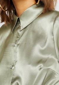 Kvinna i en ljusgrön satinskjorta med knappar, spetsig krage och stora guldörhängen.