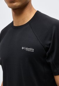 Κοντινό πλάνο ατόμου που φοράει μαύρο κοντομάνικο μπλουζάκι Columbia Titanium, εμφανίζοντας την υφή του υφάσματος και το λογότυπο της μάρκας στο στήθος.