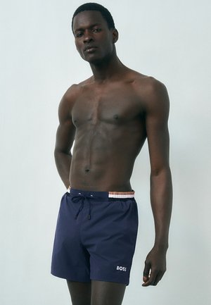 Muž bez trička v námornícky modrých plavkových šortkách BOSS so šnúrkou, stojaci na jednoduchom svetlom pozadí.