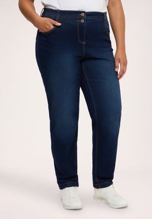 Kvinde iført mørkeblå jeans med dobbelte knapper og hvide sneakers, stående med den ene hånd i lommen mod en ensfarvet baggrund.