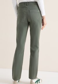 Personne portant un pantalon droit vert olive, un haut blanc et des baskets blanches avec des rayures vertes, se tenant devant un fond uni.