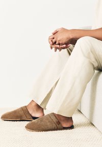 Braune gewebte Hausschuhe mit weicher dunkler Innenausstattung, einem Schlupfdesign und flacher Sohle, auf einem strukturierten hellen Teppich platziert.