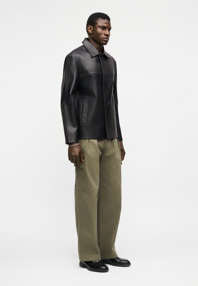 Άνδρας με μαύρο δερμάτινο μπουφάν, ελαιδί παντελόνι cargo, μαύρα παπούτσια και ασημένιο βραχιόλι, που στέκεται μπροστά σε απλό λευκό φόντο.