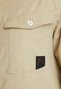 Beige Hemd mit einer Klapptasche auf der Brust, einem weißen Knopf und einem schwarzen Patch mit orangefarbenen Nähten, aus leichtem Stoff gefertigt.