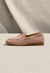 Ροζ δερμάτινο μοκασίνι με ξύλινη σόλα και χρυσά ρουρικά, παρουσιάζεται μπροστά σε ουδέτερο υφασμάτινο φόντο.