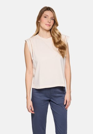 Joven mujer con cabello largo y ondulado que lleva una camiseta blanca sin mangas con detalles recortados y pantalones azul oscuro, de pie frente a un fondo liso.