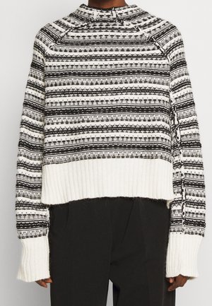 Pull tricoté à rayures horizontales noires et blanches, avec un ourlet et des poignets blancs côtelés épais, porté avec un pantalon noir.