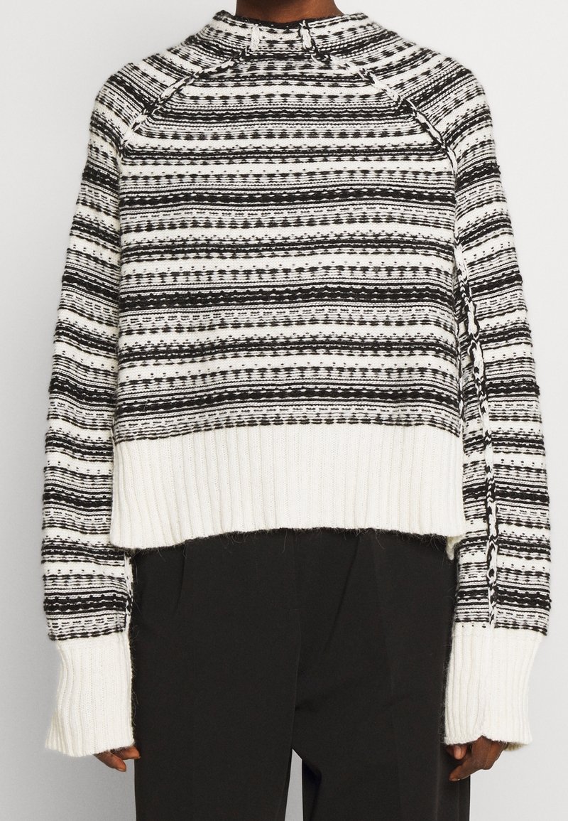 Pull tricoté à rayures horizontales noires et blanches, avec un ourlet et des poignets blancs côtelés épais, porté avec un pantalon noir.