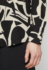 Svart og kremfarget bluse med abstrakt geometrisk mønster, glatt tekstur, detaljer med knappede mansjetter og løs passform, vist med en hånd på ermet.
