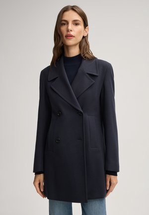 Femme portant un manteau doublé bleu marine foncé sur un col roulé noir avec un jean bleu clair sur un fond uni.