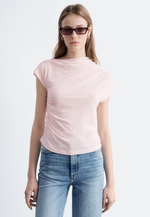 Young woman wearing light pink ruched top, blue jeans, dark rectangular sunglasses, and silver hoop earrings against a white background.