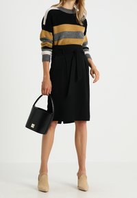 Suéter a rayas en negro, amarillo y gris; falda negra con cinturón; bolso tipo cubo negro texturizado; tacones beige. Estilo minimalista y líneas limpias.