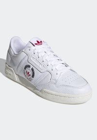 Sneaker bianco in pelle con punta perforata, lati testurizzati e un logo rosso sulla linguetta. Presenta dettagli di cucitura e suola in gomma bianca.