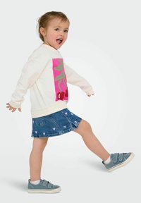 Pull blanc avec un graphique de cerises roses et vertes, jupe en denim bleu avec des motifs de cœurs, et baskets à velcro grises, dans une pose ludique.