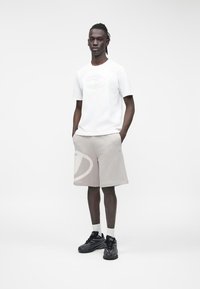 Moški stoji, oblečen v belo kratko srajco z reliefnim vzorcem, bež kratke hlače z velikim logotipom, bele nogavice in črne superge, roke v žepih.