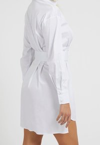 Abito camicia bianco con maniche lunghe e polsini con bottone. Presenta una vita stretta e dettagli con pieghe sul retro, realizzato in tessuto leggero.
