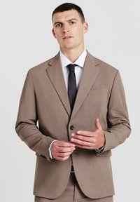 Hellbraunes Sakko mit Revers, einziger Knopfverschluss und einer Brusttasche. Getragen über einem weißen Hemd und einer schwarzen Krawatte.