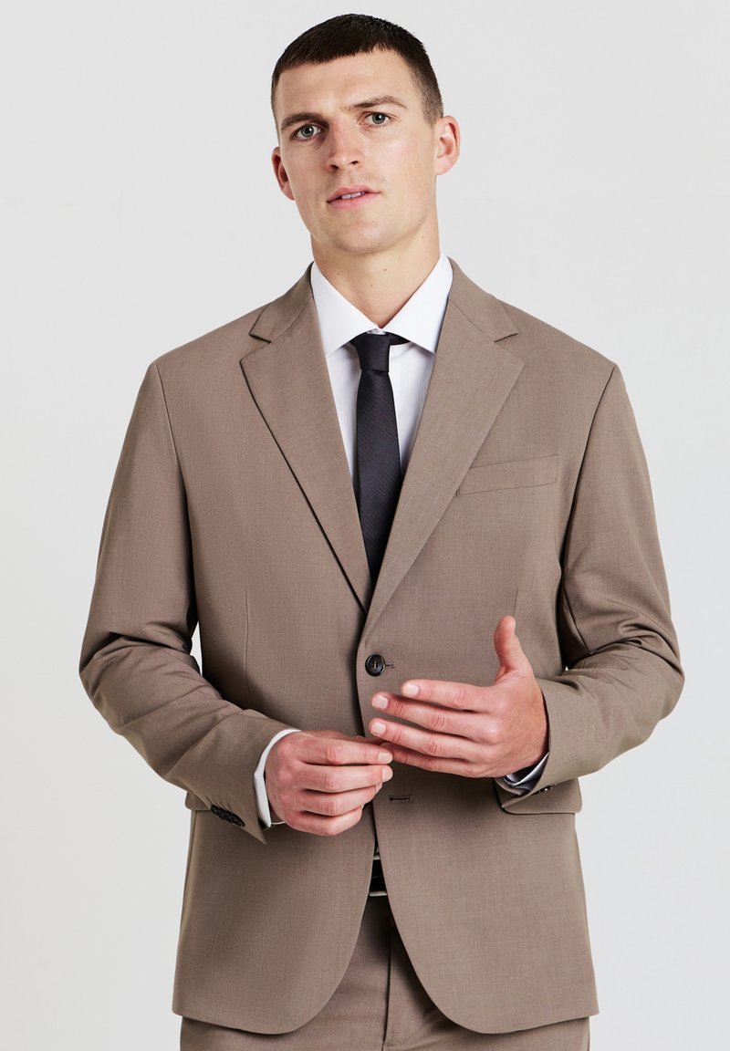 Hellbraunes Sakko mit Revers, einziger Knopfverschluss und einer Brusttasche. Getragen über einem weißen Hemd und einer schwarzen Krawatte.