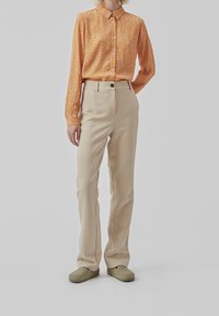 Pomarańczowa koszula z długim rękawem w biały kwiatowy wzór, zestawiona z beżowymi spodniami o wysokim stanie i prostym kroju. Model nosi oliwkowe buty typu slip-on.