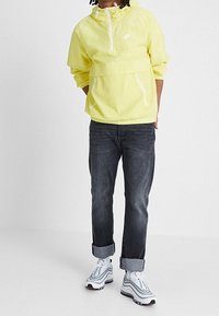 Ljusgul huvan vindjacka med framsida dragkedja och två vita dragkedjafickor, tillsammans med mörkgrå jeans med uppvikta ärmar och vita sneakers.